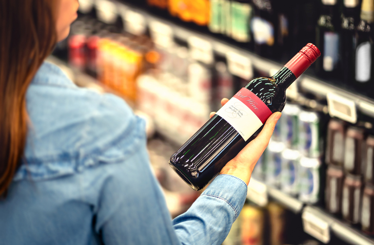 Une femme lit l'étiquette d'une bouteille de vin rouge dans un magasin de vins et spiritueux ou au rayon alcools d'un supermarché. Rayon rempli de boissons alcoolisées. Une cliente tient une bouteille de merlot et fait son choix.
